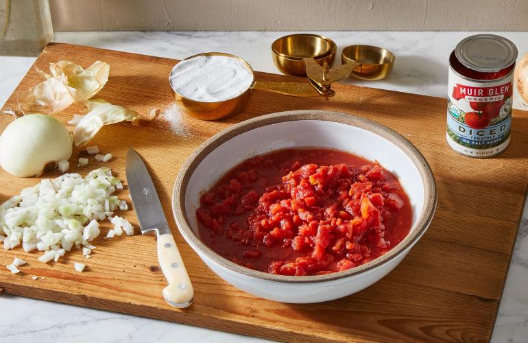 A bowl of diced tomatoes on a cutting board next to a chopped onion, salt and can of Muir Glen Organic Diced Tomatoes.