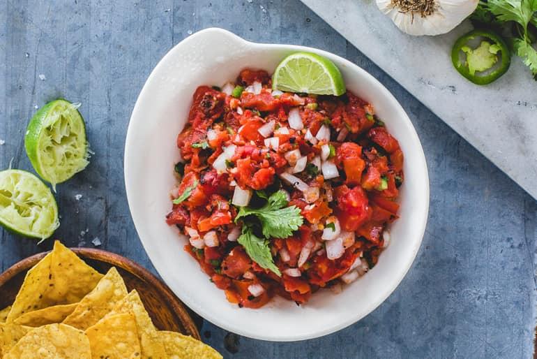 Overhead view of a bowl of salsa picada made with Muir Glen Organic Fire Roasted Diced Tomatoes garnished with cilantro and lime, served with yellow corn chips.