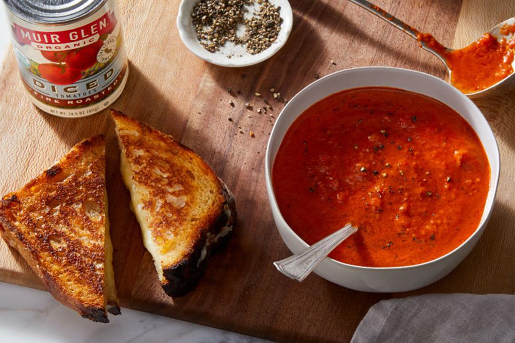 A bowl of tomato soup and a grilled cheese next to a can of Muir Glen Organic Diced Tomatoes on a wood cutting board.