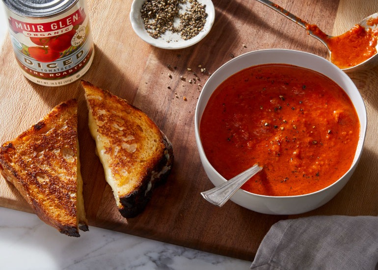 A bowl of tomato soup and a grilled cheese next to a can of Muir Glen Organic Diced Tomatoes on a wood cutting board.